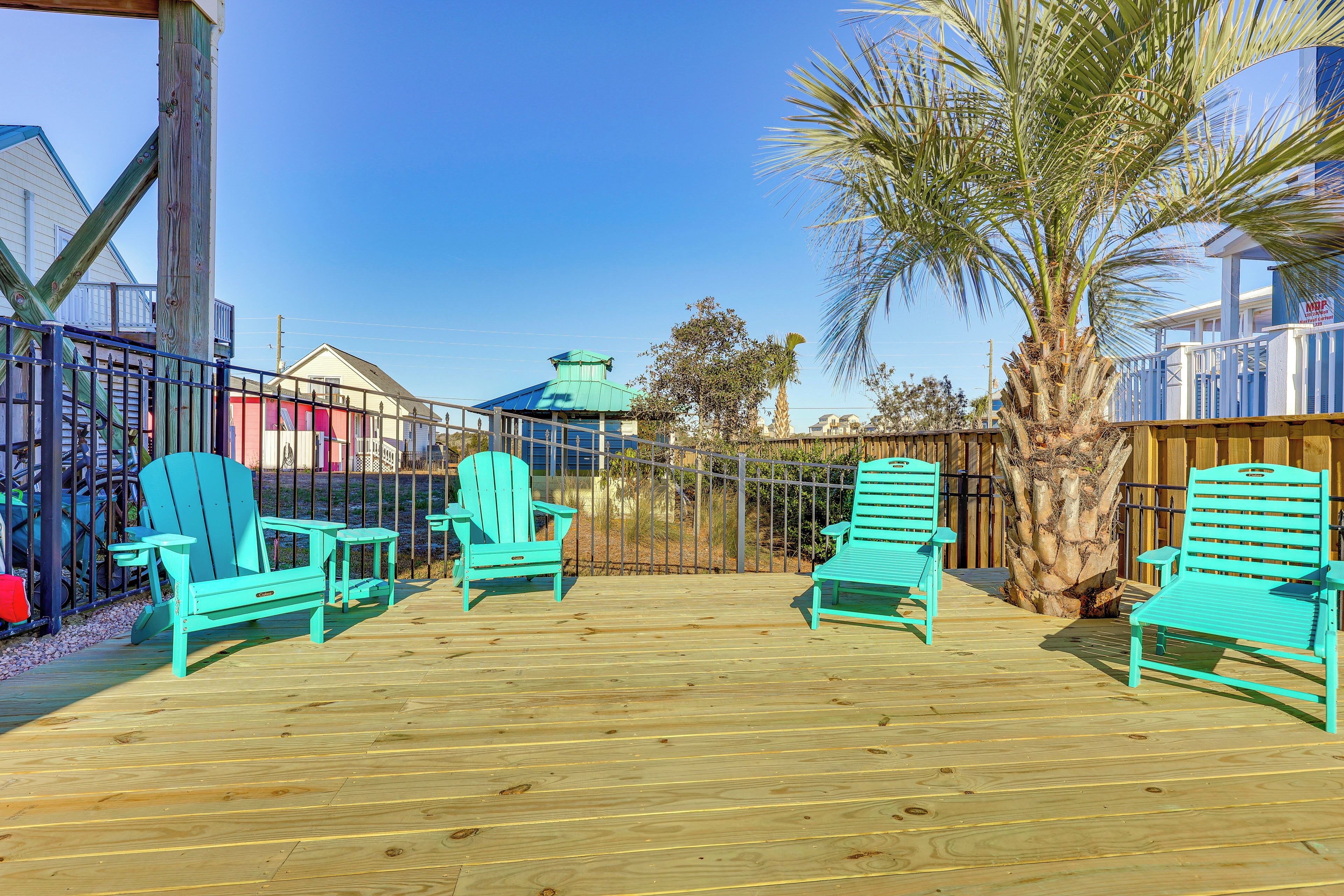 A wooden deck with turquoise Adirondack chairs, a palm tree, and a blue-roofed building in the background.