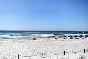 Ein Strand mit weißem Sand, blauem Himmel und dem Meer im Hintergrund. Entlang der Küste stehen blaue Sonnenschirme und Liegestühle.