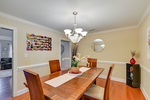 Une salle à manger avec une table en bois, des chaises, un lustre, un vase rempli de fleurs et une œuvre d’art colorée accrochée au mur.