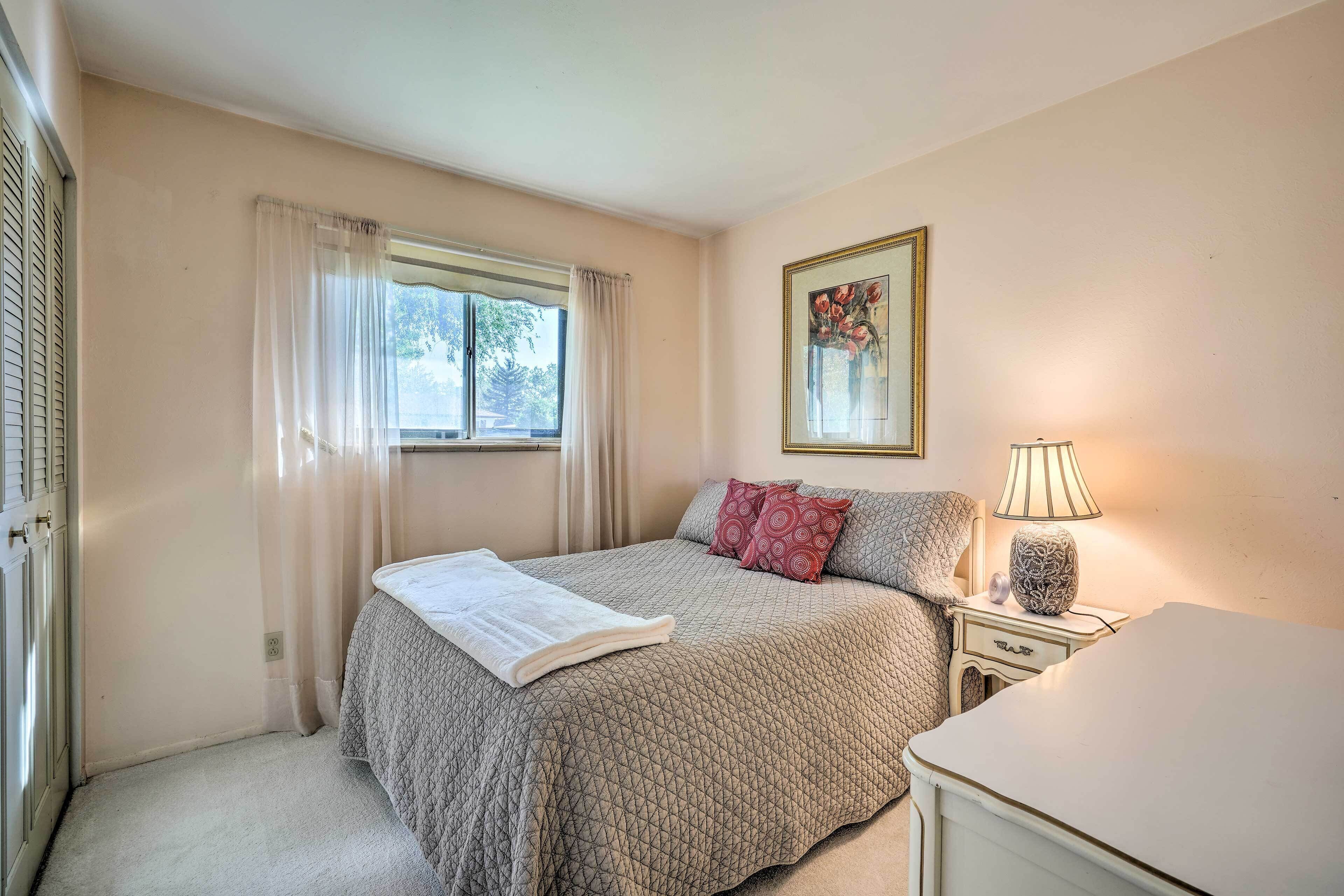 Un dormitorio con cama, mesita de noche con lámpara, ventana con cortinas e imagen enmarcada en la pared.