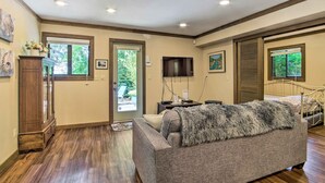 A spacious living room with a large gray sofa, wooden flooring, a wooden cabinet, a television, and a bed with a metal frame. - Coos Bay Cottage w/ Fireplace & BBQ Patio! (Coos Bay)