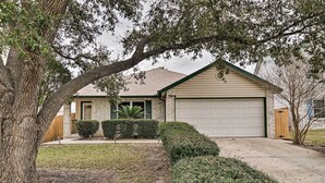 A single-story house with a garage, a front yard with a tree and shrubs, and a brick exterior. - Dog-friendly Texas Retreat w/ Hot Tub, Patio & Bar (Converse)