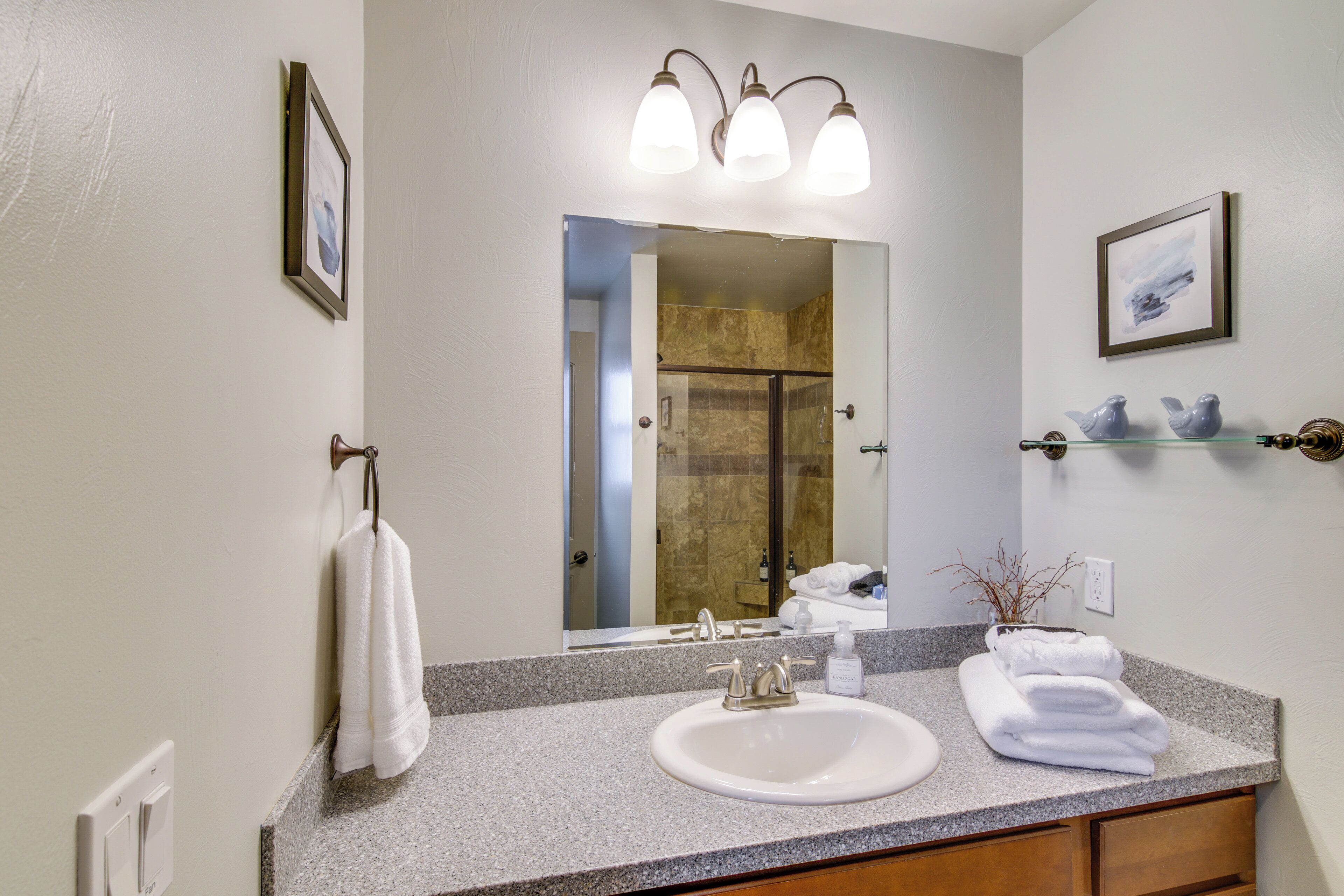 A bathroom with a granite countertop, a white sink, a mirror with three lights, a towel rack, a framed picture, and a small shelf with decorative items.