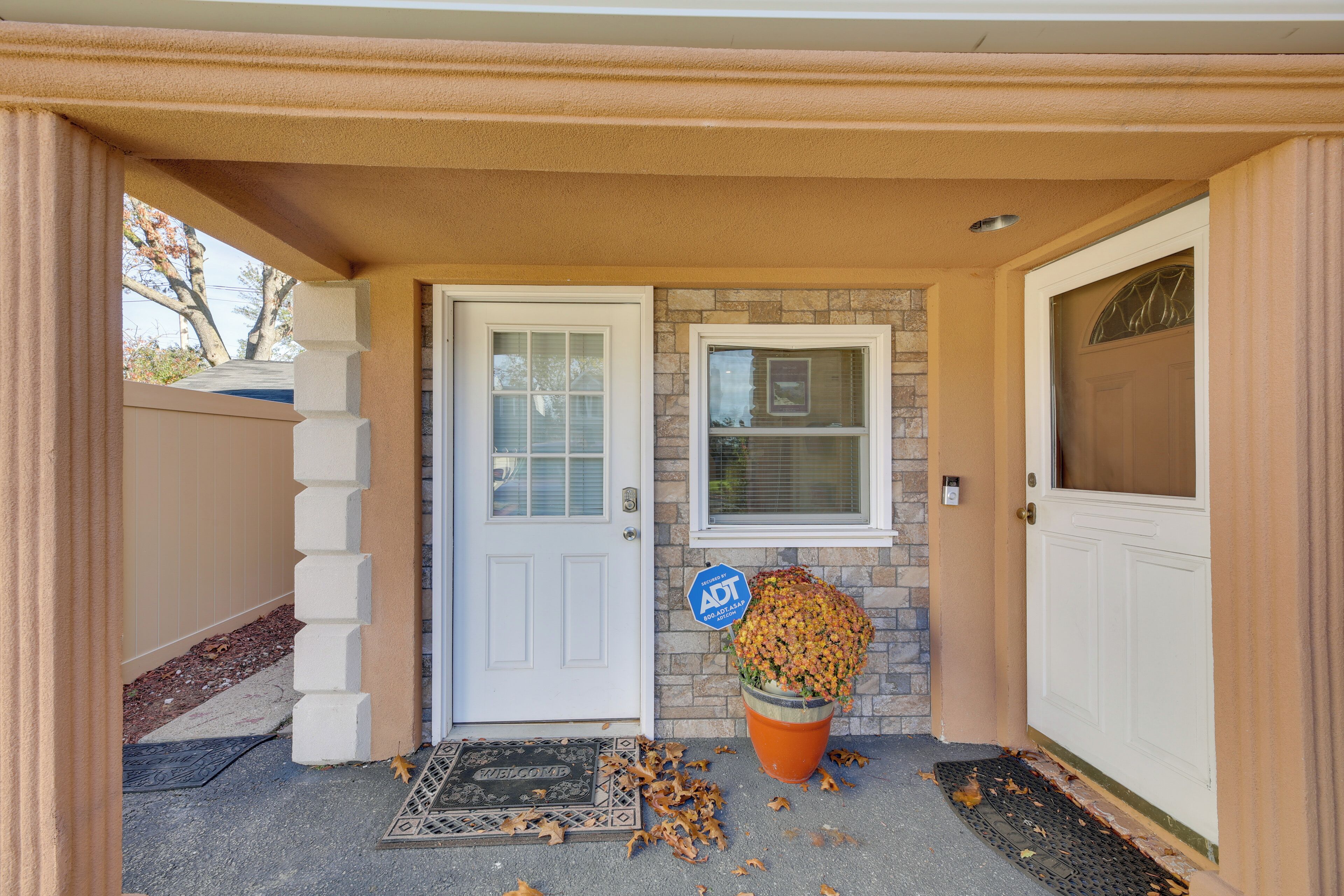 A house entrance with a white door, a welcome mat, a potted plant, and a blue sign.