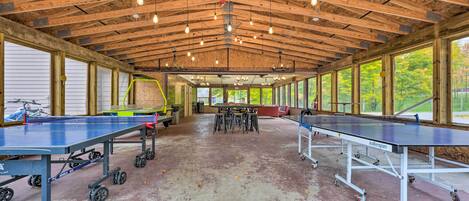 A spacious indoor area with two ping pong tables, a wooden ceiling with exposed beams, and large windows offering views of greenery outside.