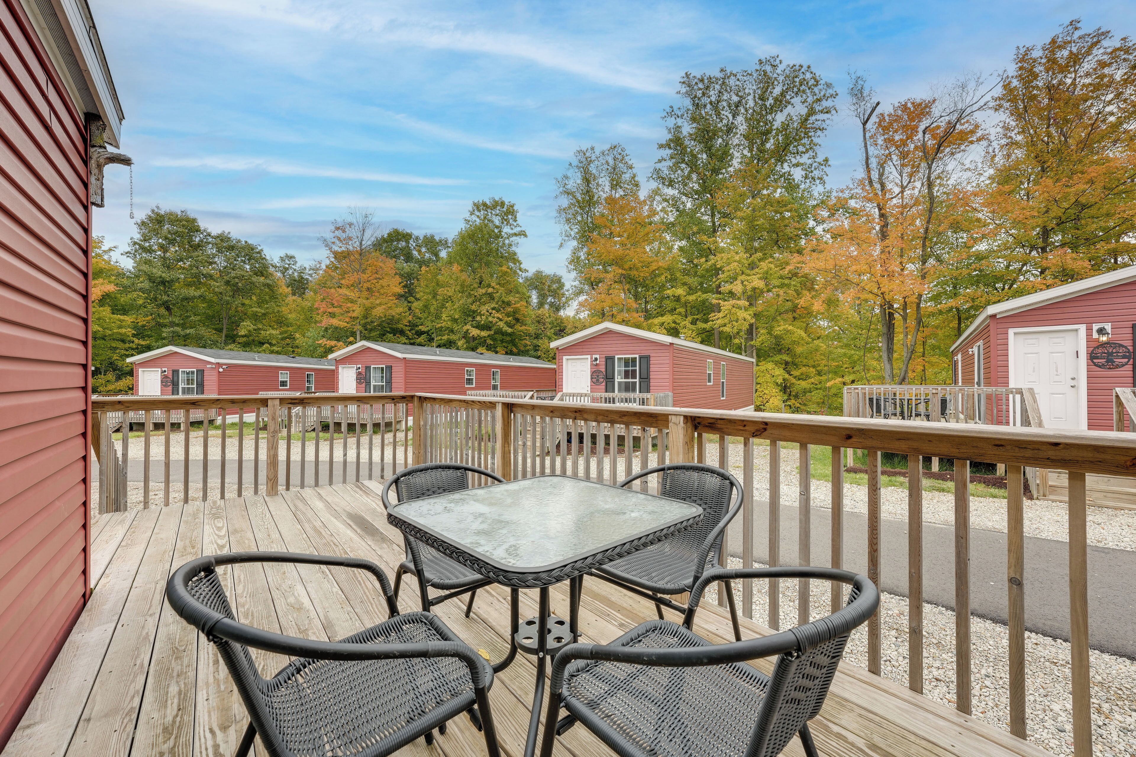 A wooden deck with a glass table and four chairs, overlooking a row of red cabins with white doors.