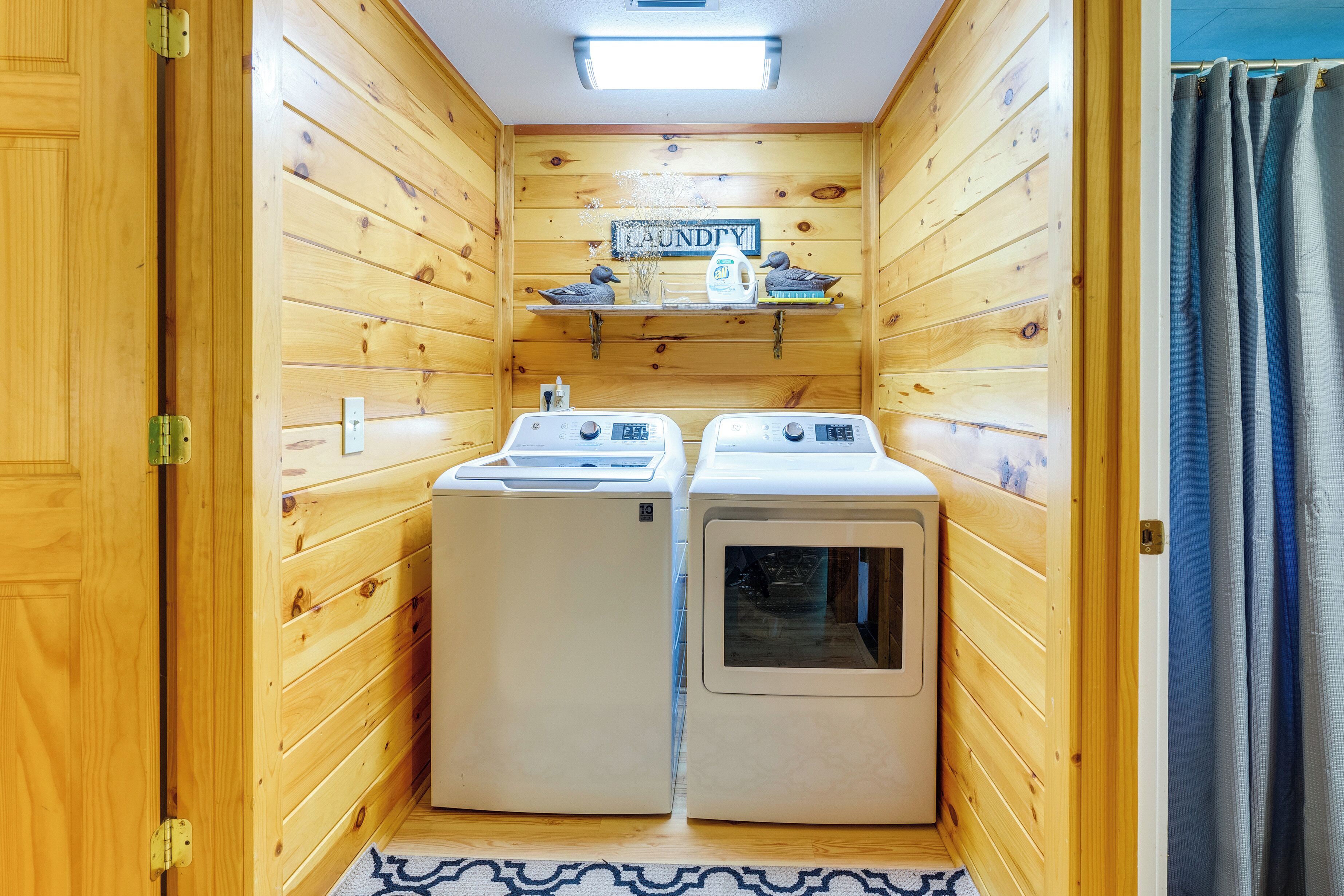 Une buanderie avec des murs en bois, deux machines à laver, une étagère remplie de produits de lessive et un panneau indiquant « Buanderie ».