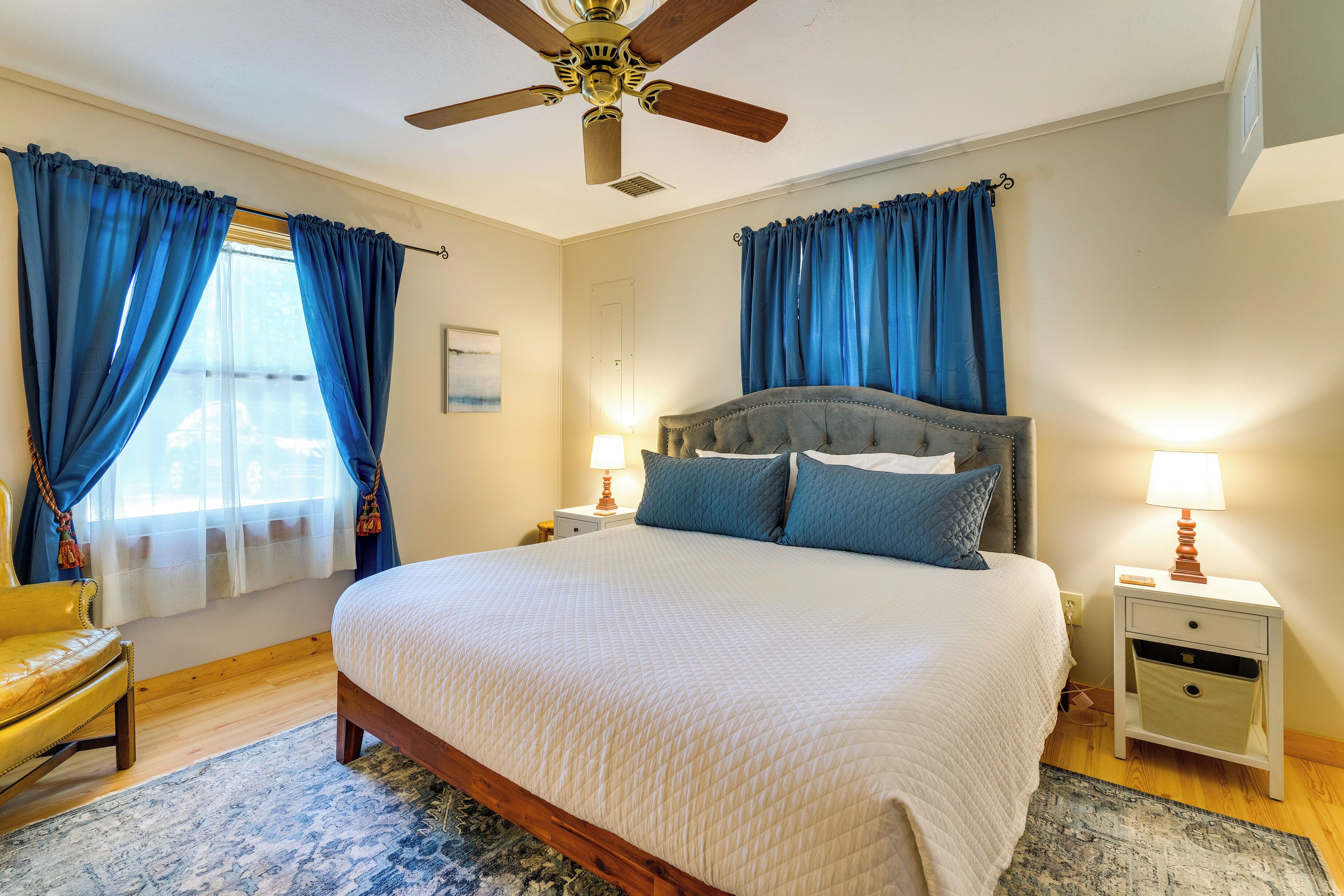 A bedroom with a large bed, two bedside tables with lamps, a ceiling fan, and a window with blue curtains.