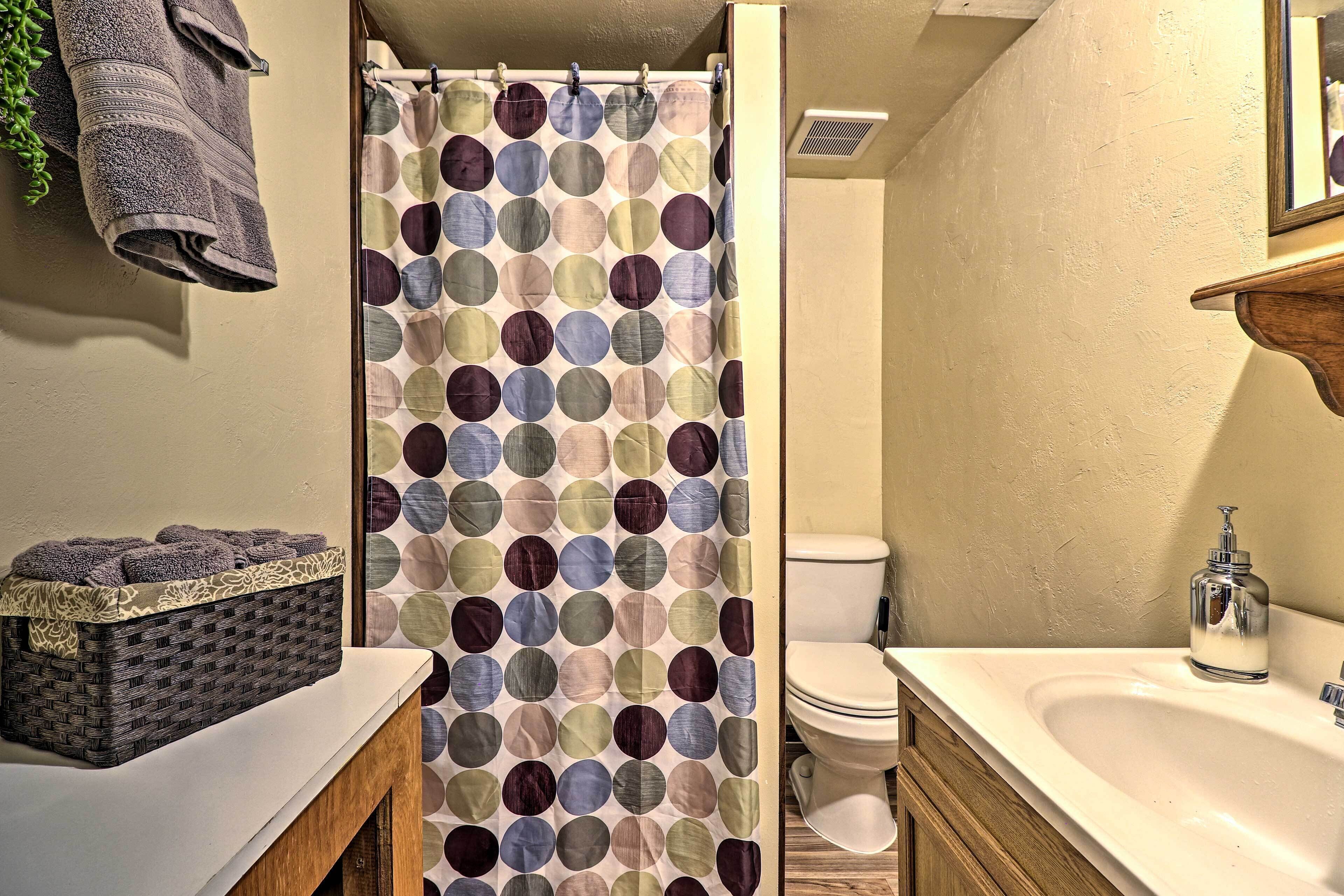A bathroom with a shower curtain, a toilet, a sink with a soap dispenser, and a basket of towels.