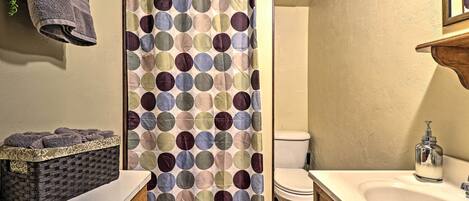 A bathroom with a shower curtain, a toilet, a sink with a soap dispenser, and a basket of towels.