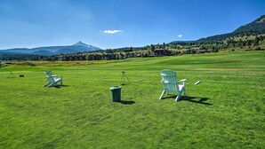 A grassy field with two white chairs and a trash bin, a mountain in the background, and a clear sky. - Big Sky Condo w/ Resort Amenities & Mountain Views (Big Sky)