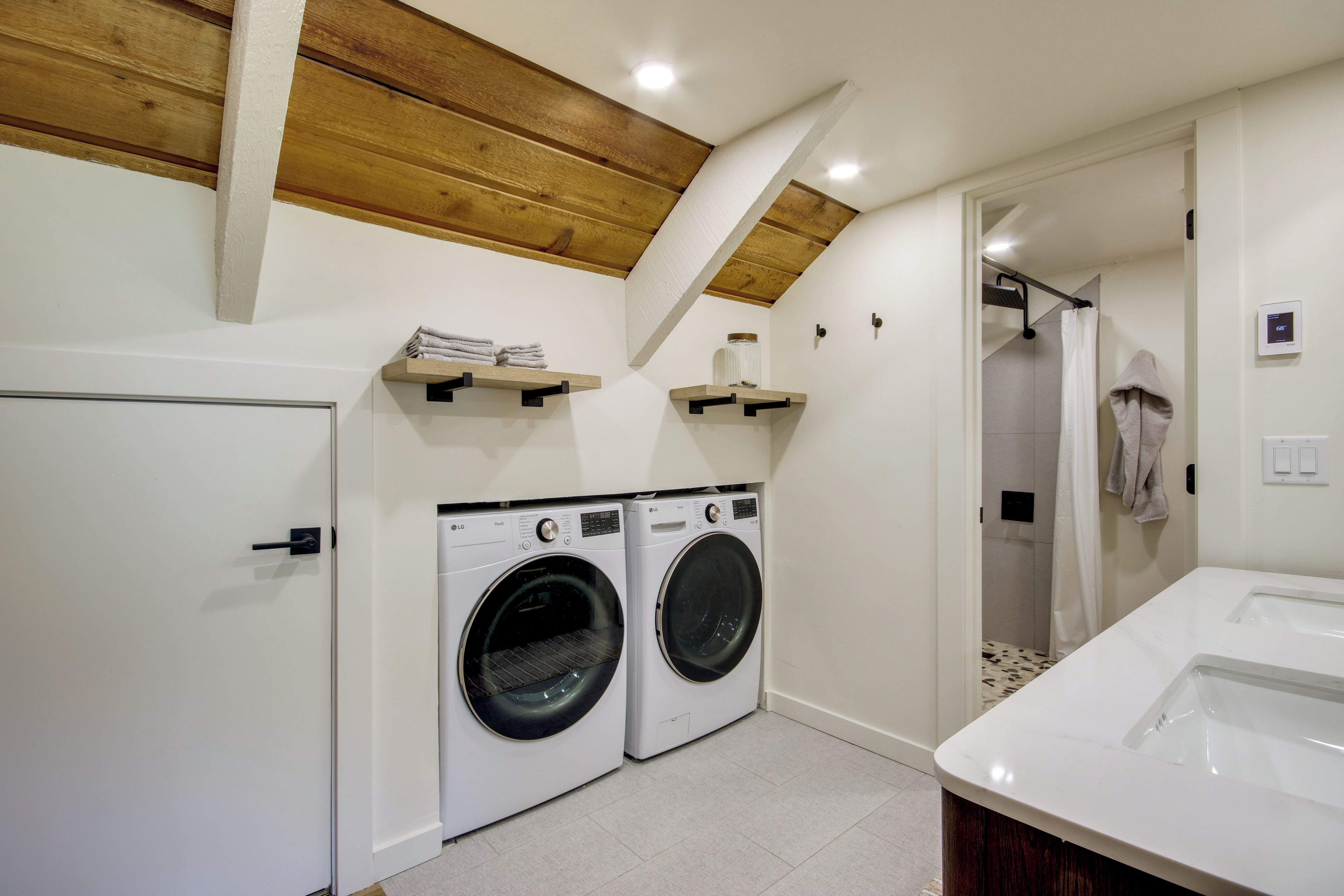 A laundry room with two front-loading washing machines, a wooden ceiling, and a shower area.