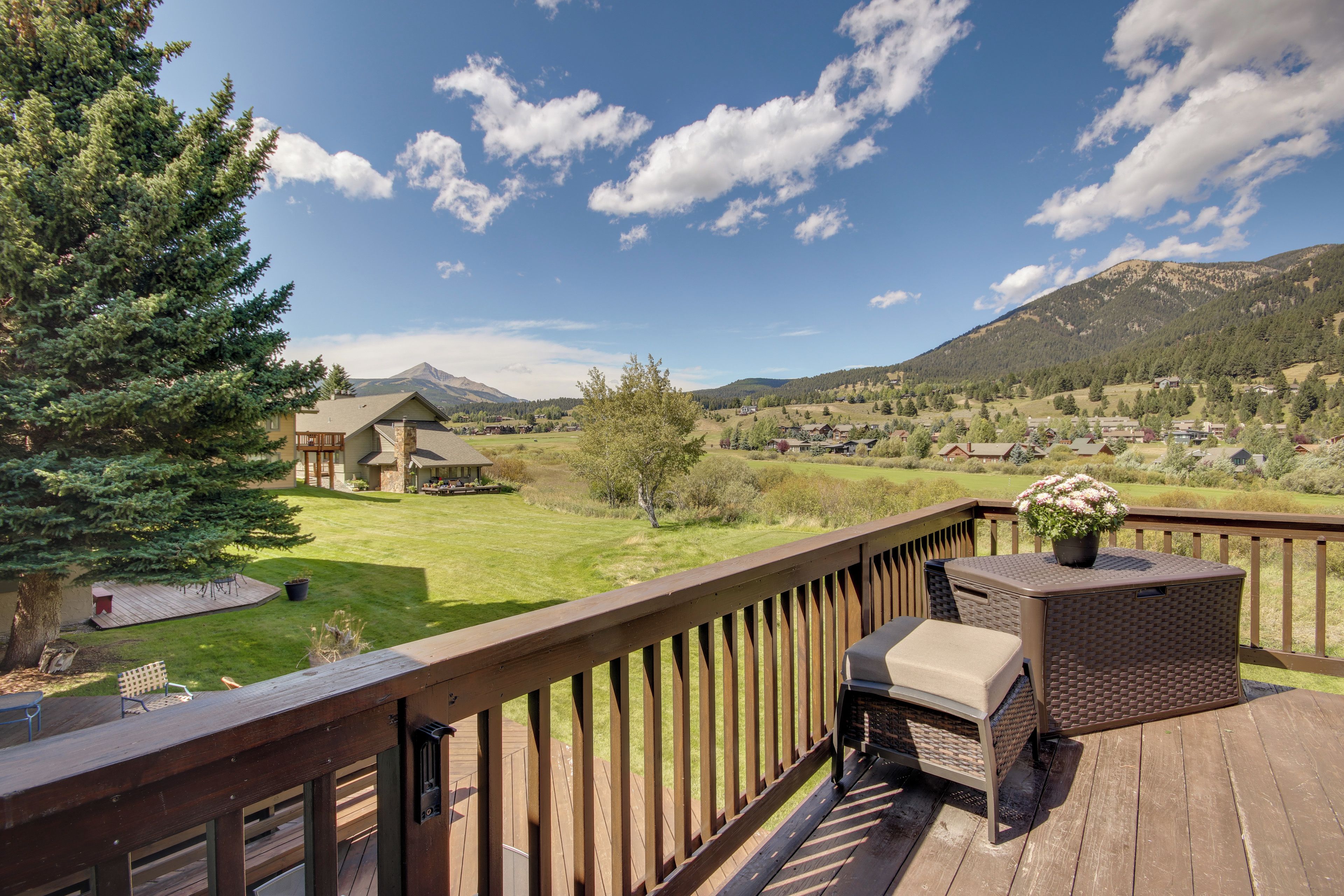 A wooden deck with outdoor furniture, overlooking a green landscape with houses and mountains.