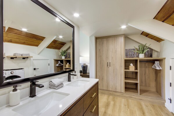 Een moderne badkamer met een grote spiegel, dubbele wastafels, houten kastjes en een plafond met zichtbare balken.