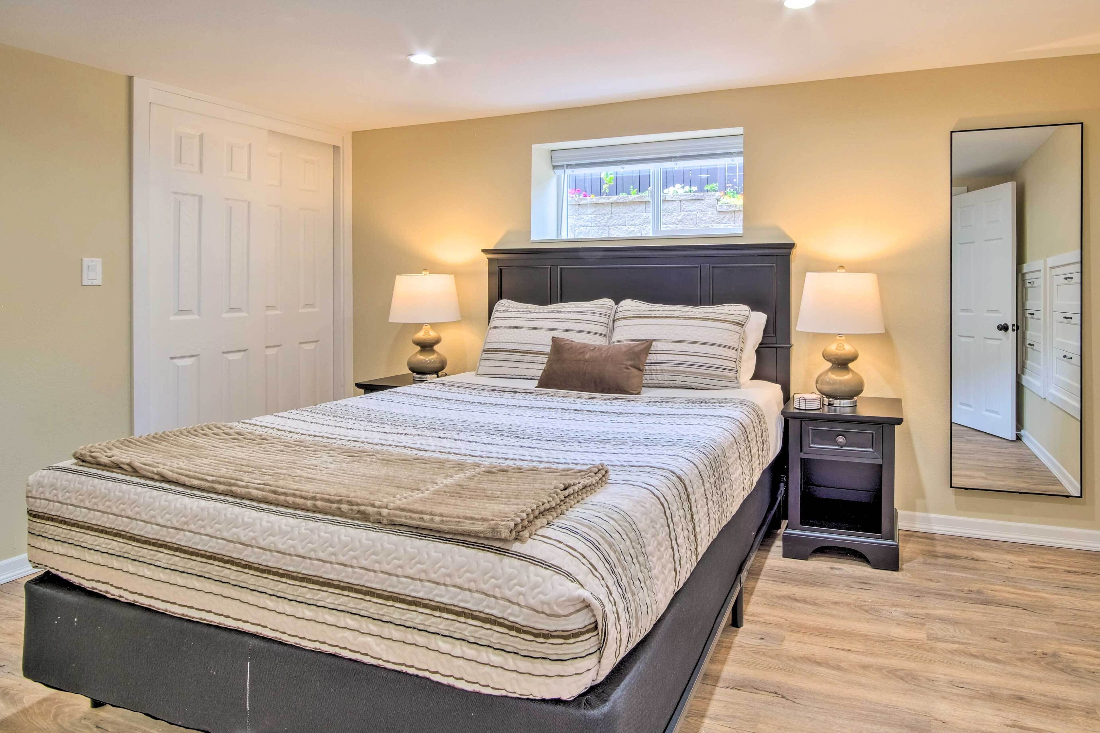 A bedroom with a large bed, two bedside tables with lamps, a window with a view of plants, and a mirror on the wall.
