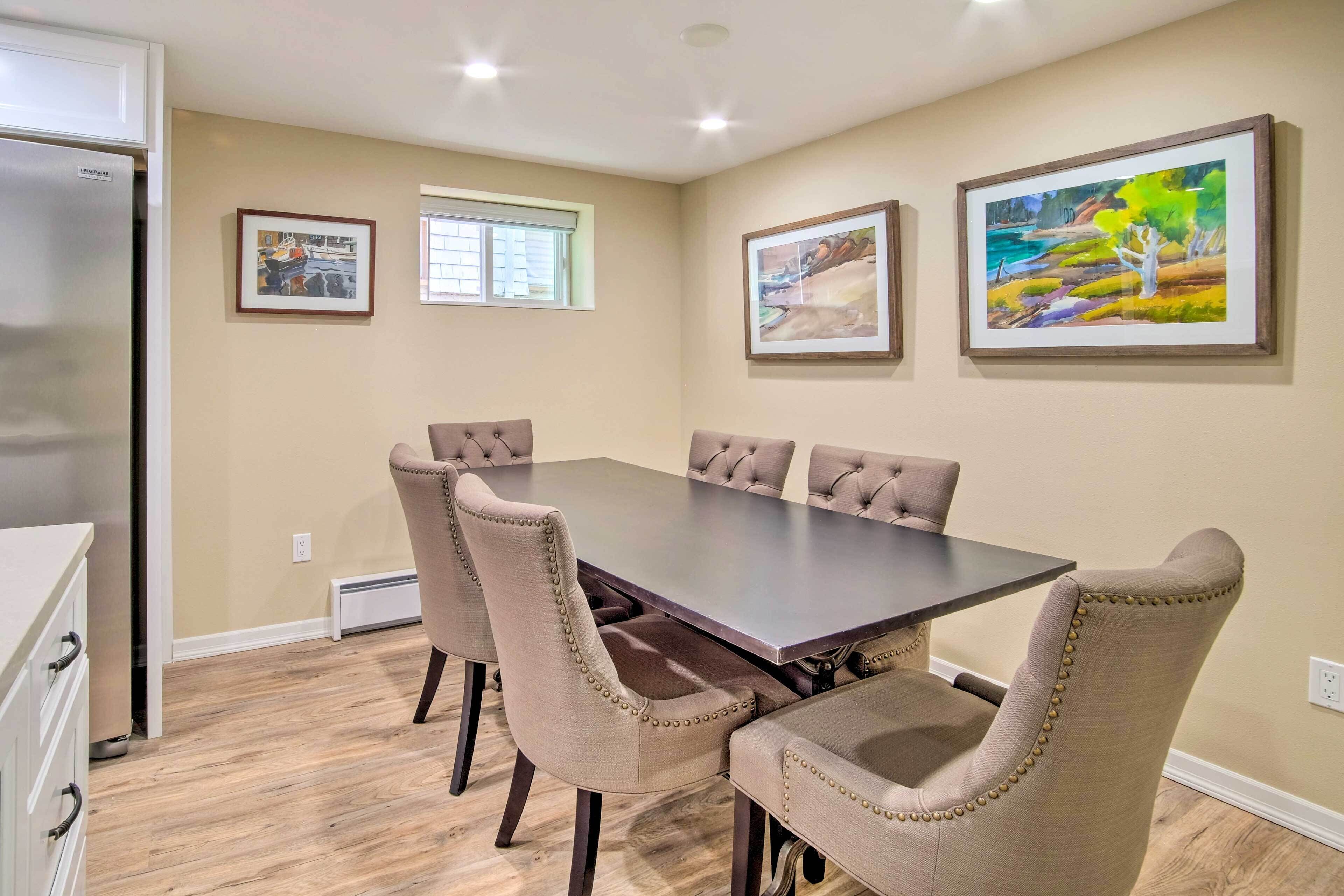 A dining room with a large table and six upholstered chairs, beige walls, and two framed pictures.