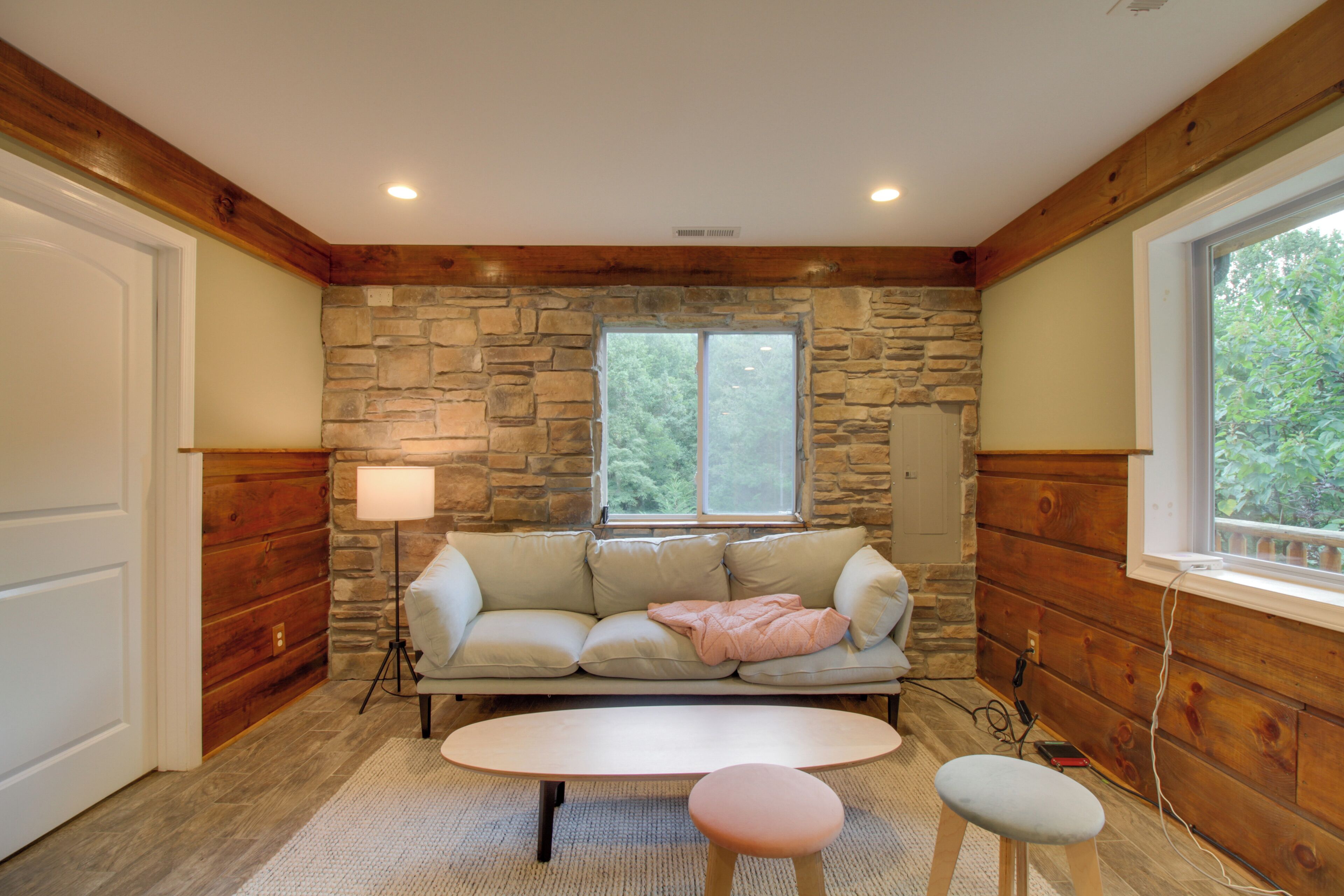 A cozy room with a stone wall, a wooden ceiling, a beige sofa, a small table, and a window with a view of greenery.