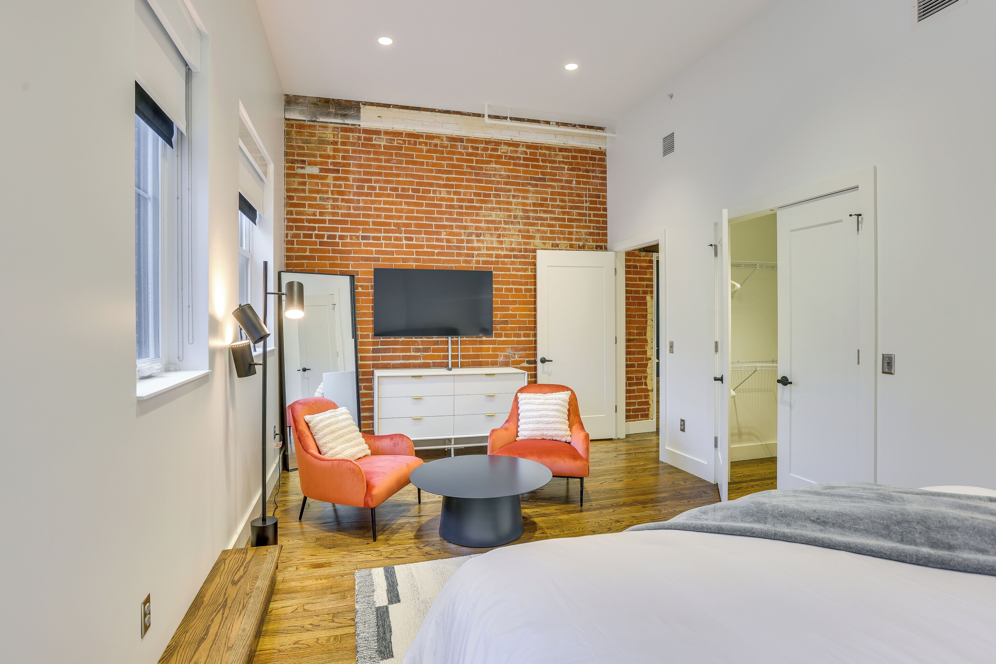 A modern bedroom with a brick wall, a flat-screen TV, two orange chairs, a round coffee table, and a bed.