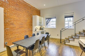 Une cuisine moderne avec un mur en briques, un coin repas avec une longue table et des chaises, et un escalier doté d’une rampe en métal.
