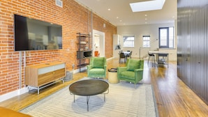 A spacious living area with a brick wall, a flat-screen TV, green armchairs, a round coffee table, and a dining area in the background. - Downtown Omaha Getaway w/ Deck & Gas Grill! (Omaha)