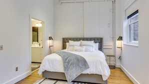A modern bedroom with a large bed, a wooden floor, and a white brick wall. - Downtown Omaha Getaway w/ Deck & Gas Grill! (Omaha)