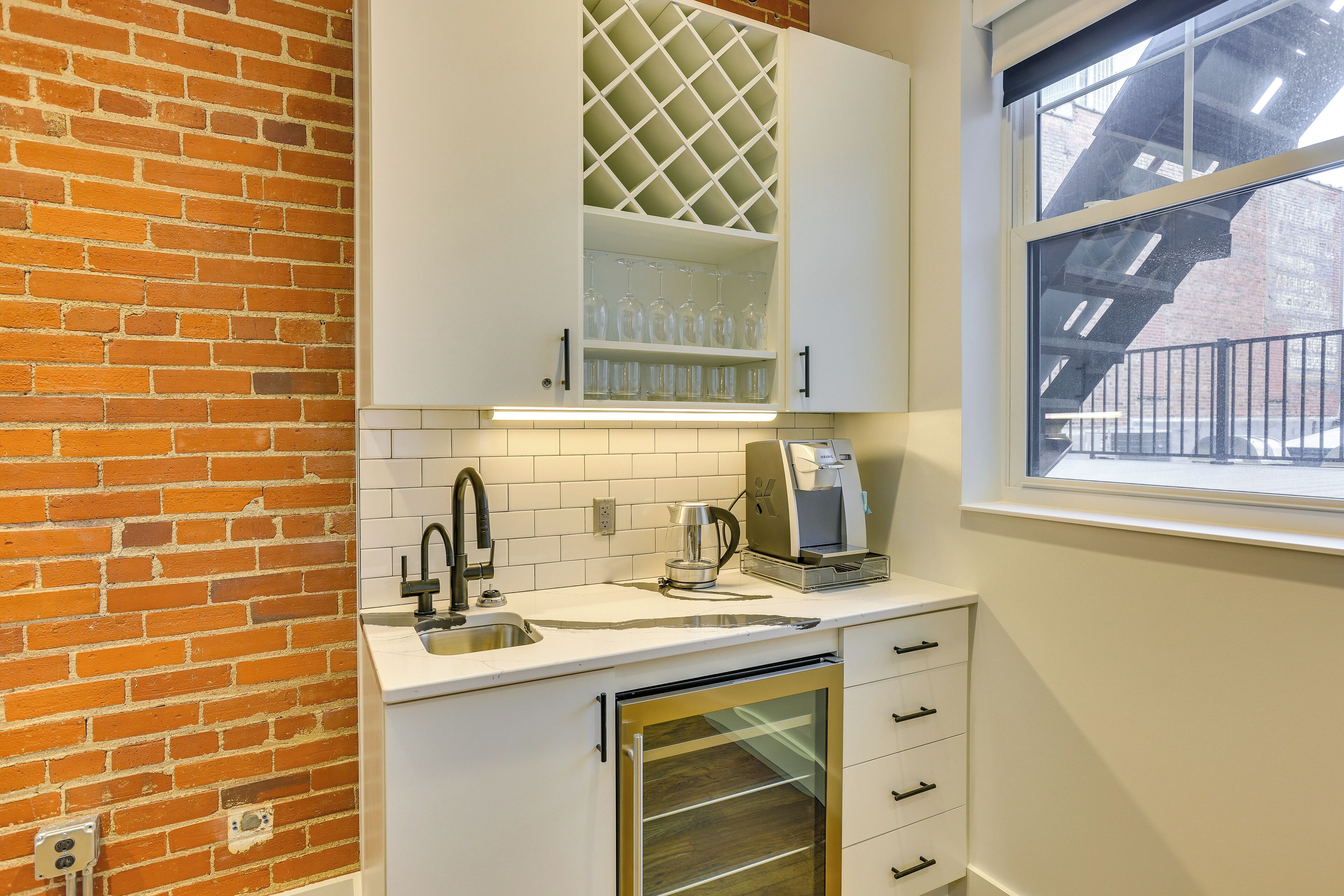 A kitchen with white cabinets, a brick wall, a window, and a dishwasher.