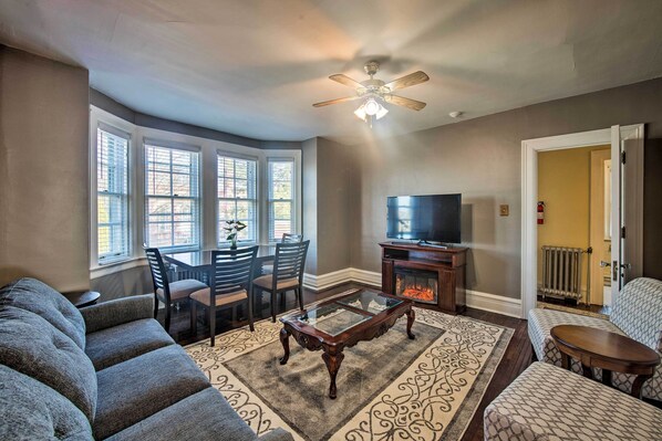 A living room with a sofa, a coffee table, a TV, and a fireplace. There are windows and a ceiling fan.