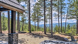 A hot tub surrounded by a stone pillar and wooden deck, set in a forested area with a view of mountains and a clear sky. - Custom Angel Fire Home: Views, Hot Tub & Fire Pit! (Angel Fire)