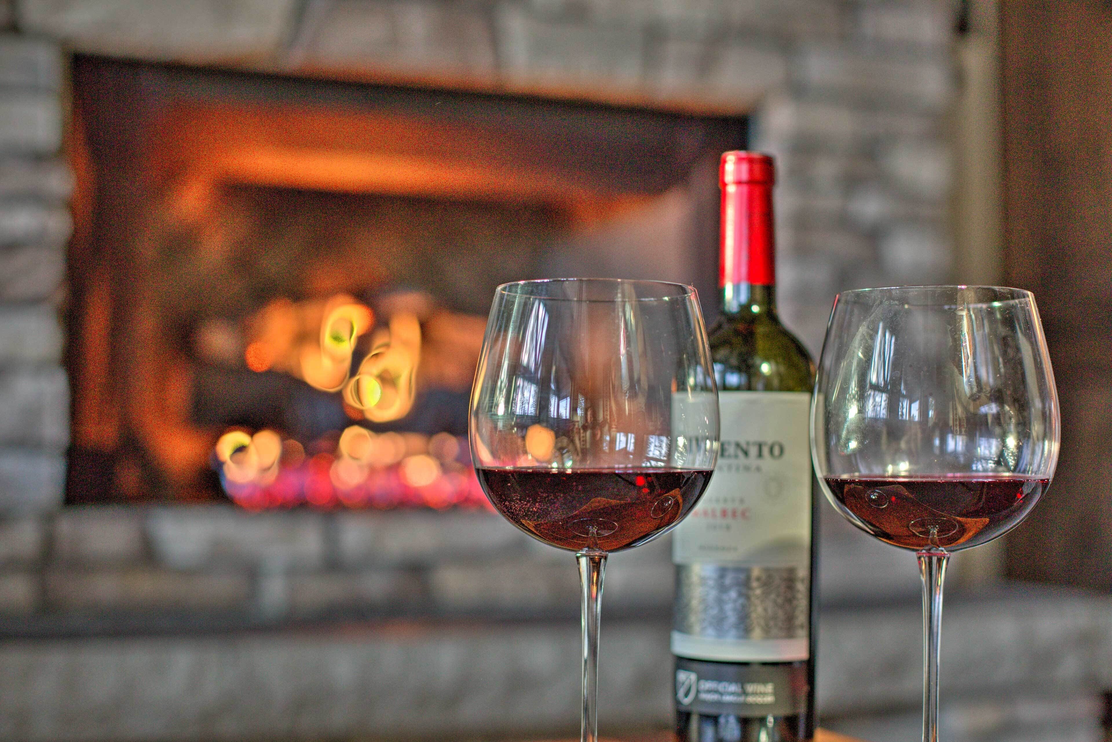 Two wine glasses with red wine in front of a fireplace and a wine bottle in the background.