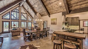 A spacious loft-style living area with a high ceiling, large windows, a fireplace, and a rustic kitchen. - Custom Angel Fire Home: Views, Hot Tub & Fire Pit! (Angel Fire)