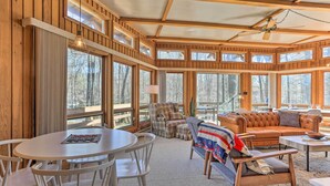 A cozy cabin interior with a round dining table, white chairs, a sofa, and a bench. Large windows offer views of the surrounding forest. - Walk to Big Bass Lake: Family Home w/ Deck! (Gouldsboro)