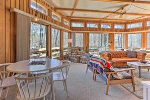 A cozy cabin interior with a round dining table, white chairs, a sofa, and a bench. Large windows offer views of the surrounding forest.
