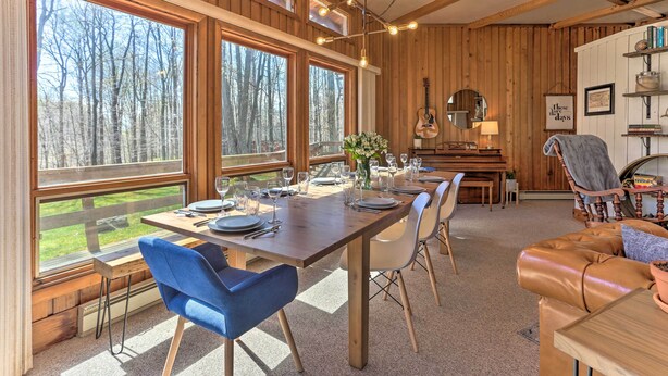 A dining area with a wooden table set for a meal, large windows with a view of trees, and a cozy seating area with a leather couch. - Walk to Big Bass Lake: Family Home w/ Deck! (Gouldsboro)