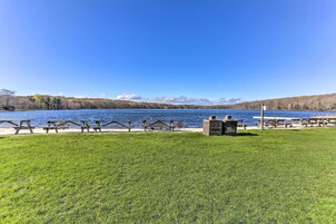 Un área de picnic junto al lago con bancos y mesas de madera, un primer plano con césped y una vista despejada del lago y montañas distantes.