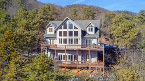 A large, two-story house with a balcony, surrounded by trees and a hilly landscape.