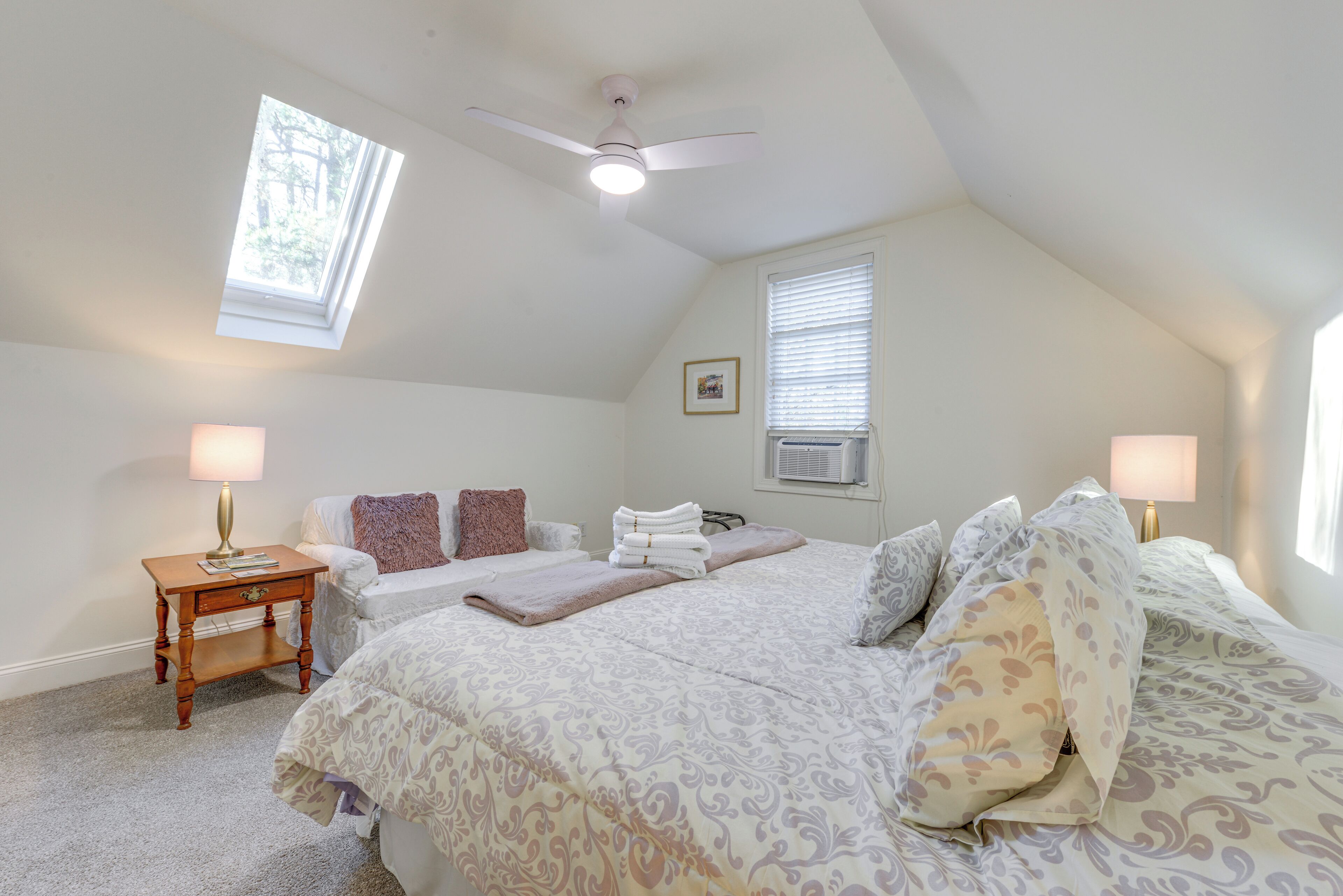 A bedroom with a sloped ceiling, a ceiling fan, a skylight, a bed with patterned bedding, two bedside tables with lamps, and an air conditioning unit.