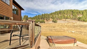 A wooden deck with a hot tub, a wooden house, and a forested hillside in the background.