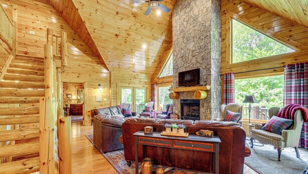 A log cabin interior with a wooden staircase, a stone fireplace, a leather sofa, and a large window overlooking greenery. - Hike, Fish & Golf: Burnsville Retreat w/ Deck! (Burnsville)