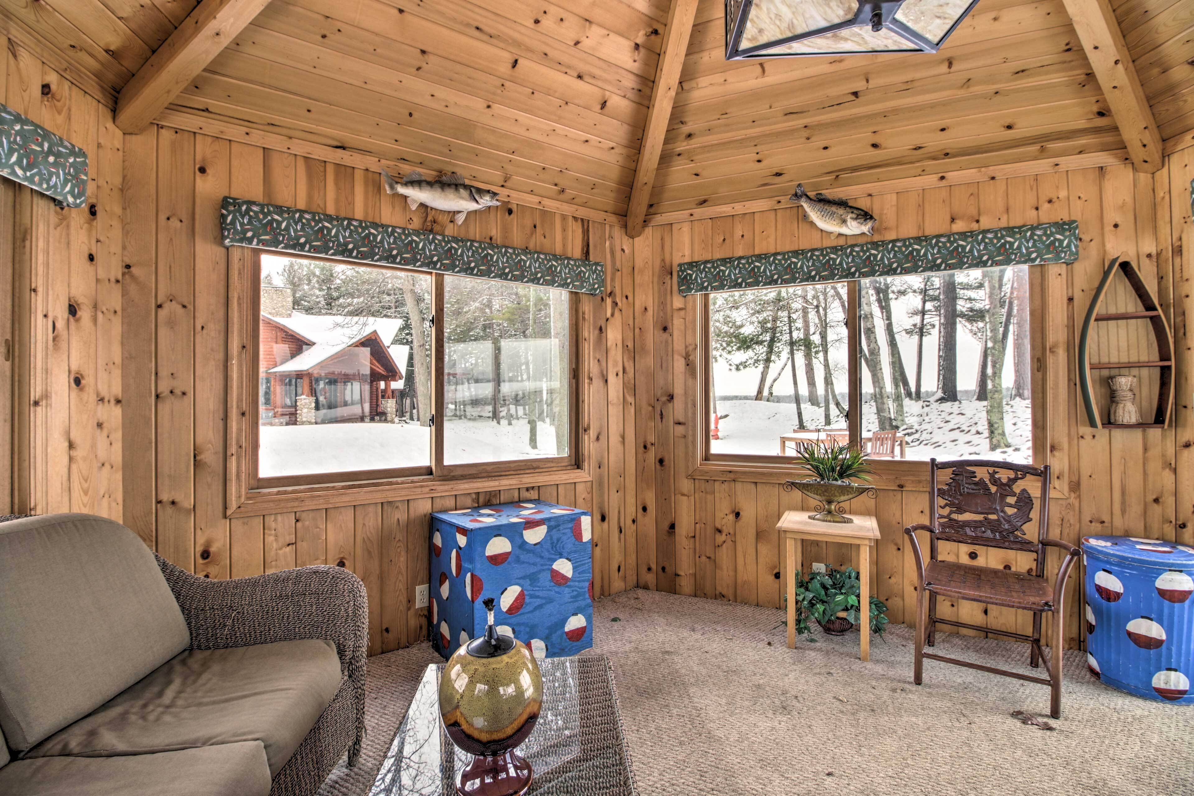 A cozy cabin interior with wooden walls, a sofa, a chair, a table with a decorative bowl, and two windows with curtains.
