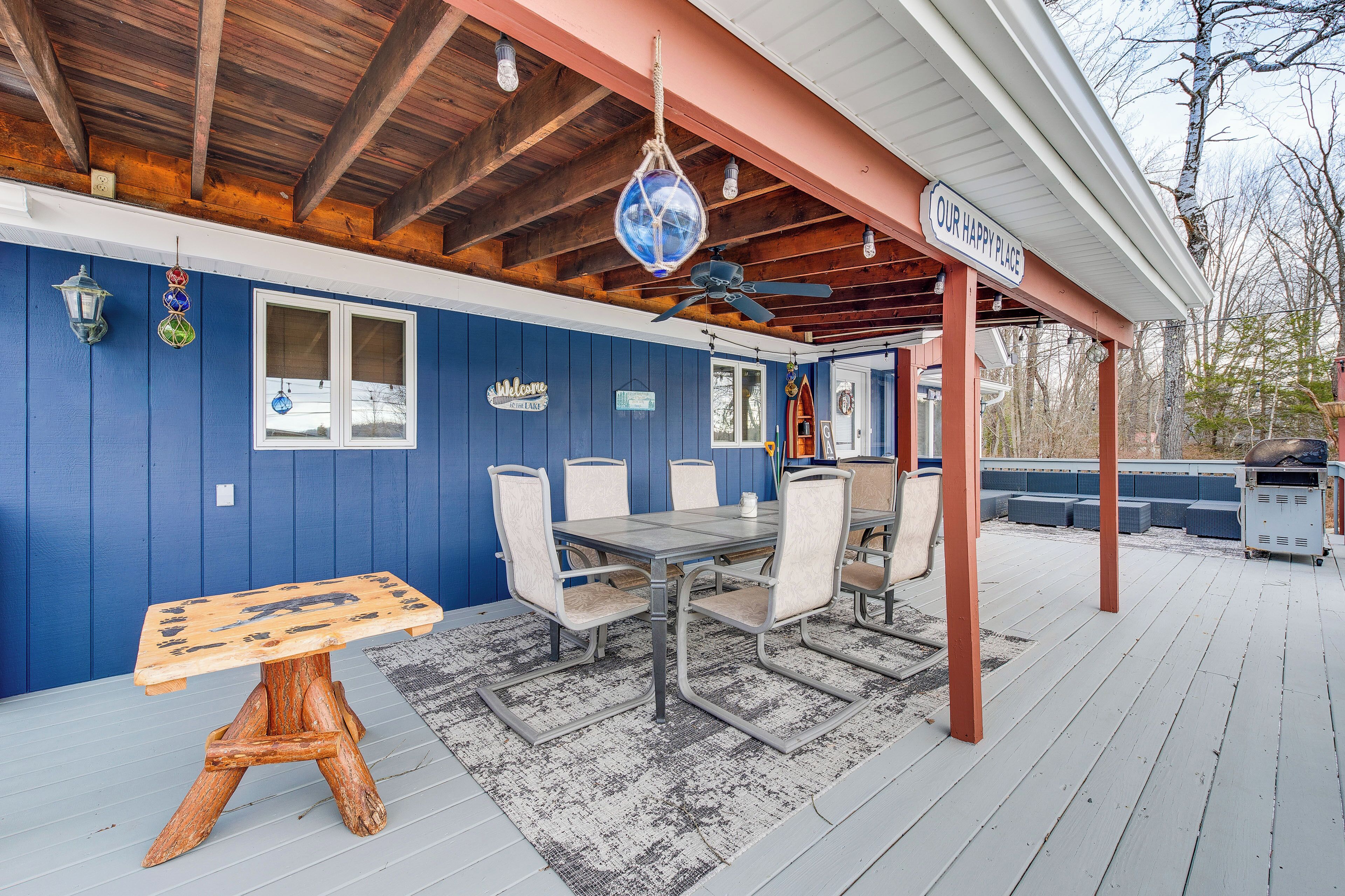 A covered outdoor patio with a wooden table and chairs, a ceiling fan, and a sign reading "OUR HAPPY PLACE".