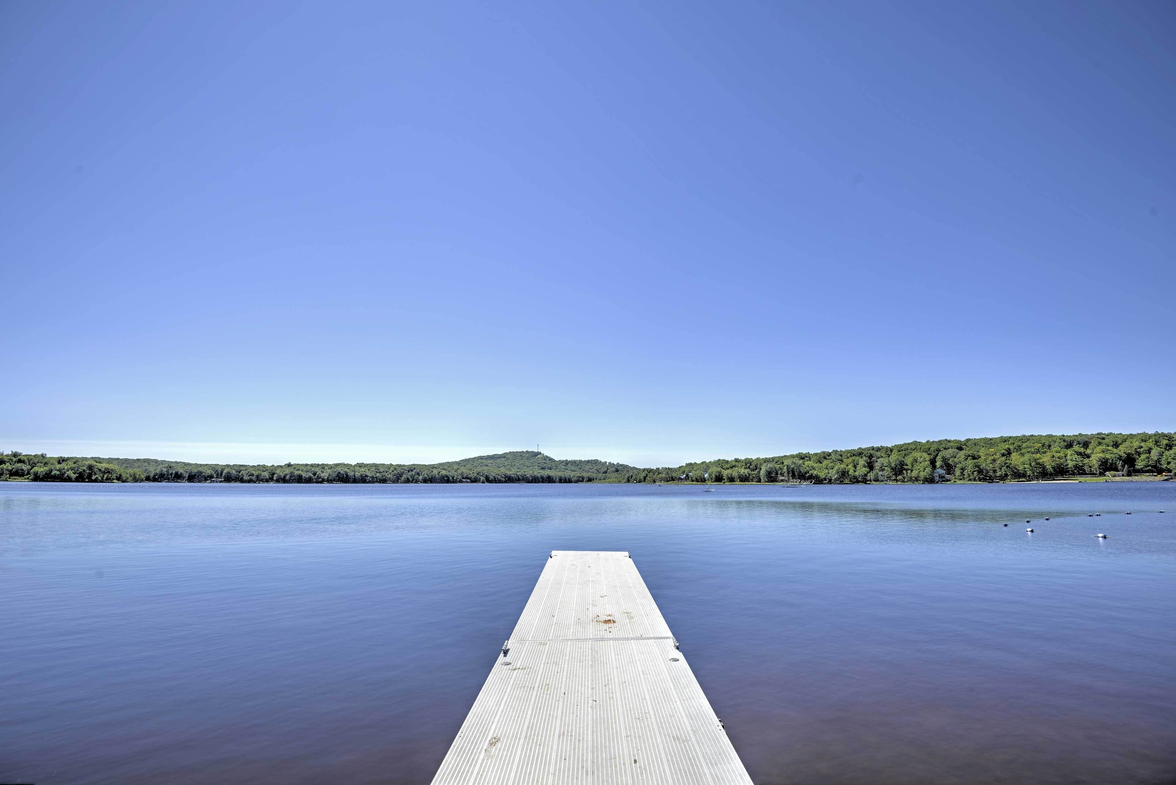 A wooden dock extending into calm water with a clear sky and a forested hill in the background.