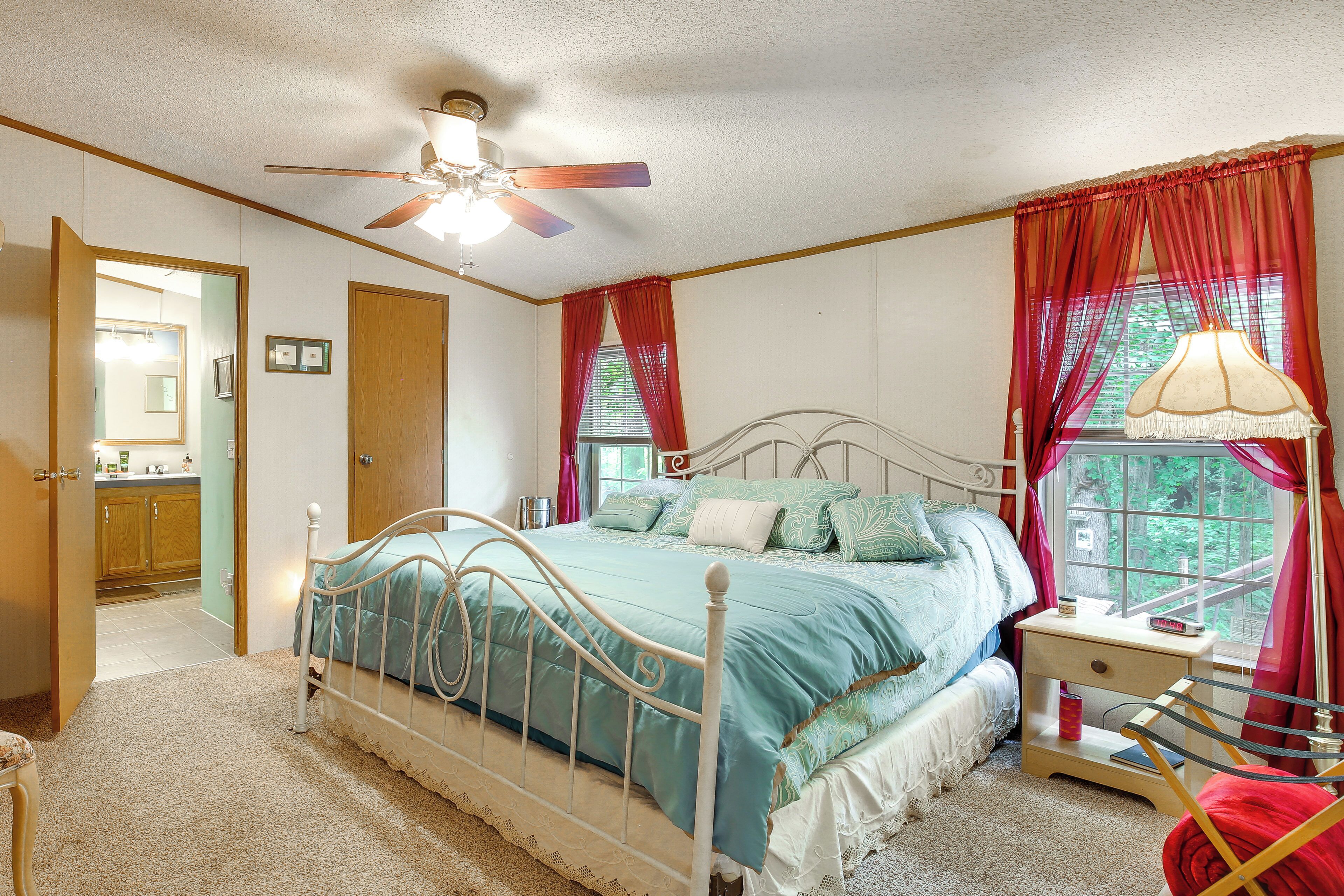 A bedroom with a bed, a nightstand, a chair, a window with curtains, a ceiling fan, and a door leading to another room.
