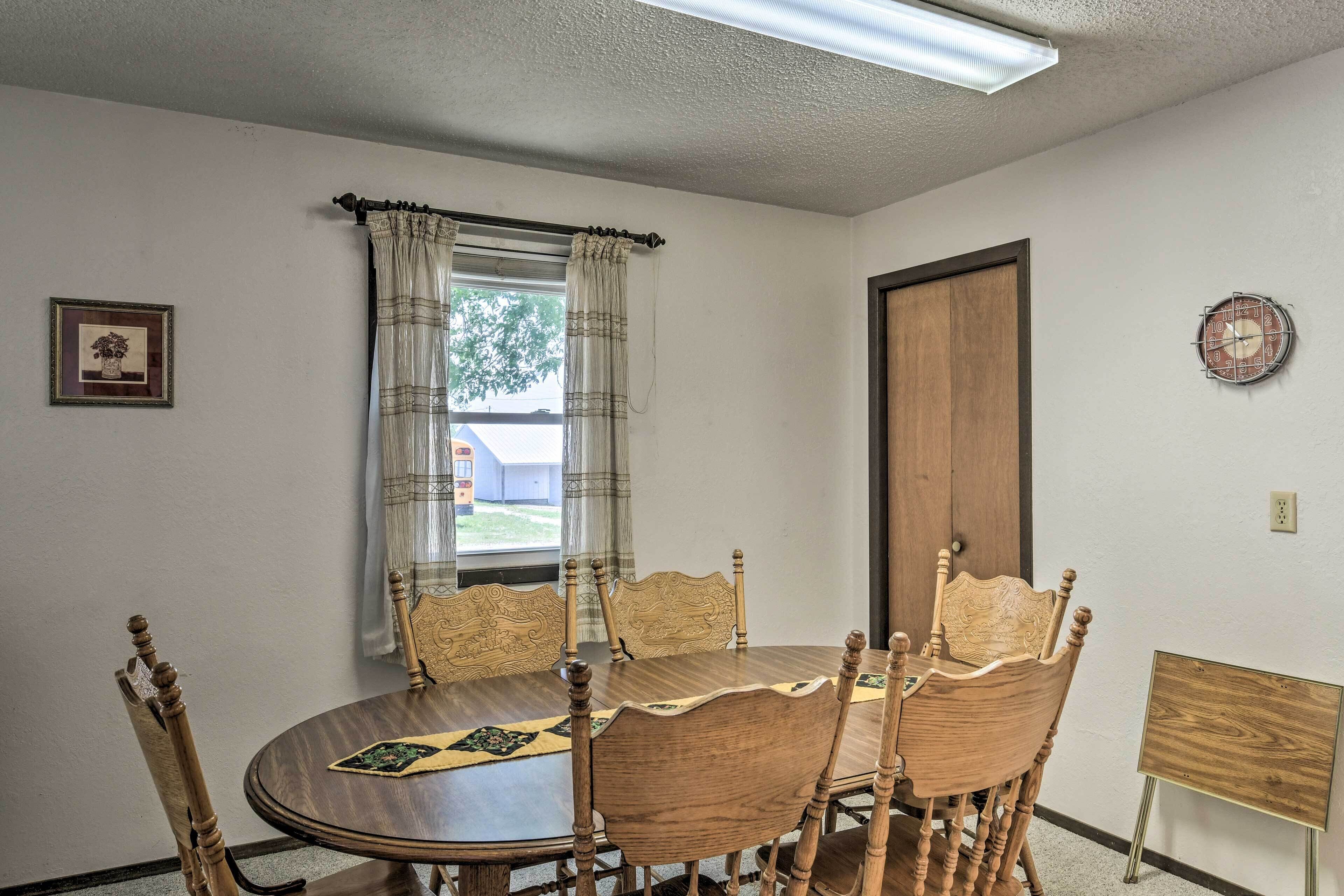 A dining area with a round wooden table, six chairs, a window with curtains, a framed picture on the wall, and a clock.