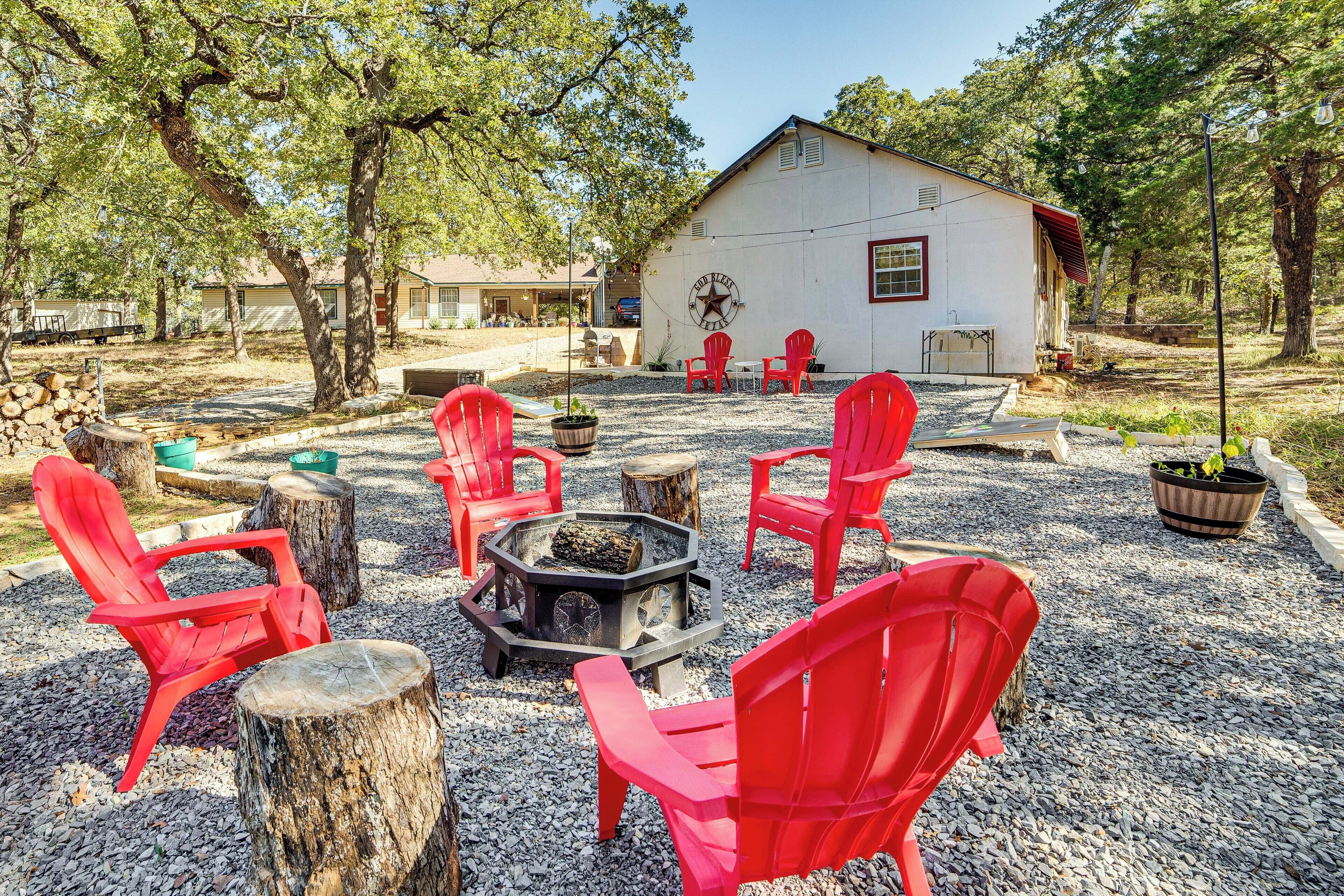 A rustic outdoor seating area with red chairs and a central fire pit, surrounded by trees and a white building in the background.