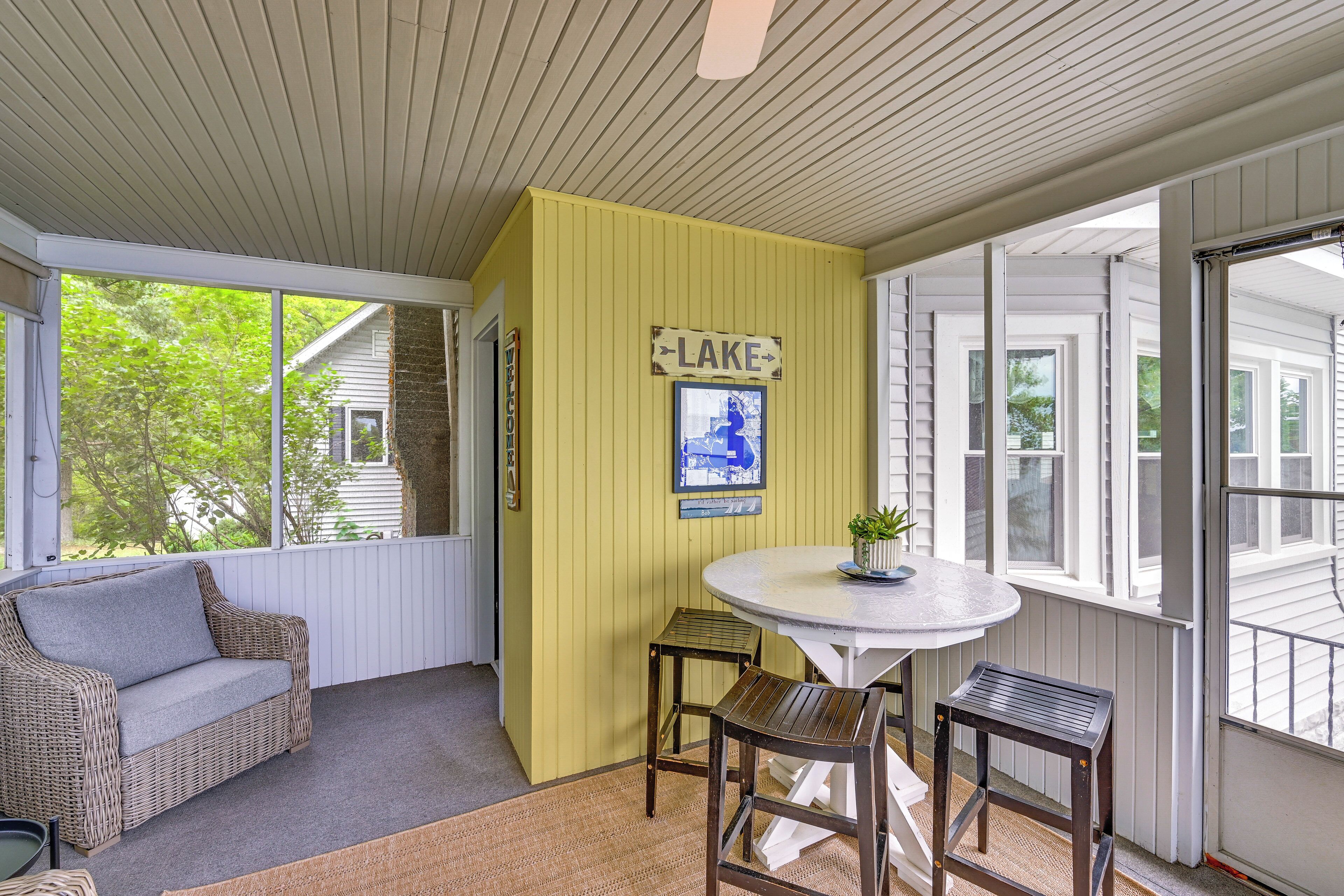 A covered porch with a wicker sofa, a small round table, and two wooden stools. A sign on the wall reads "LAKE".