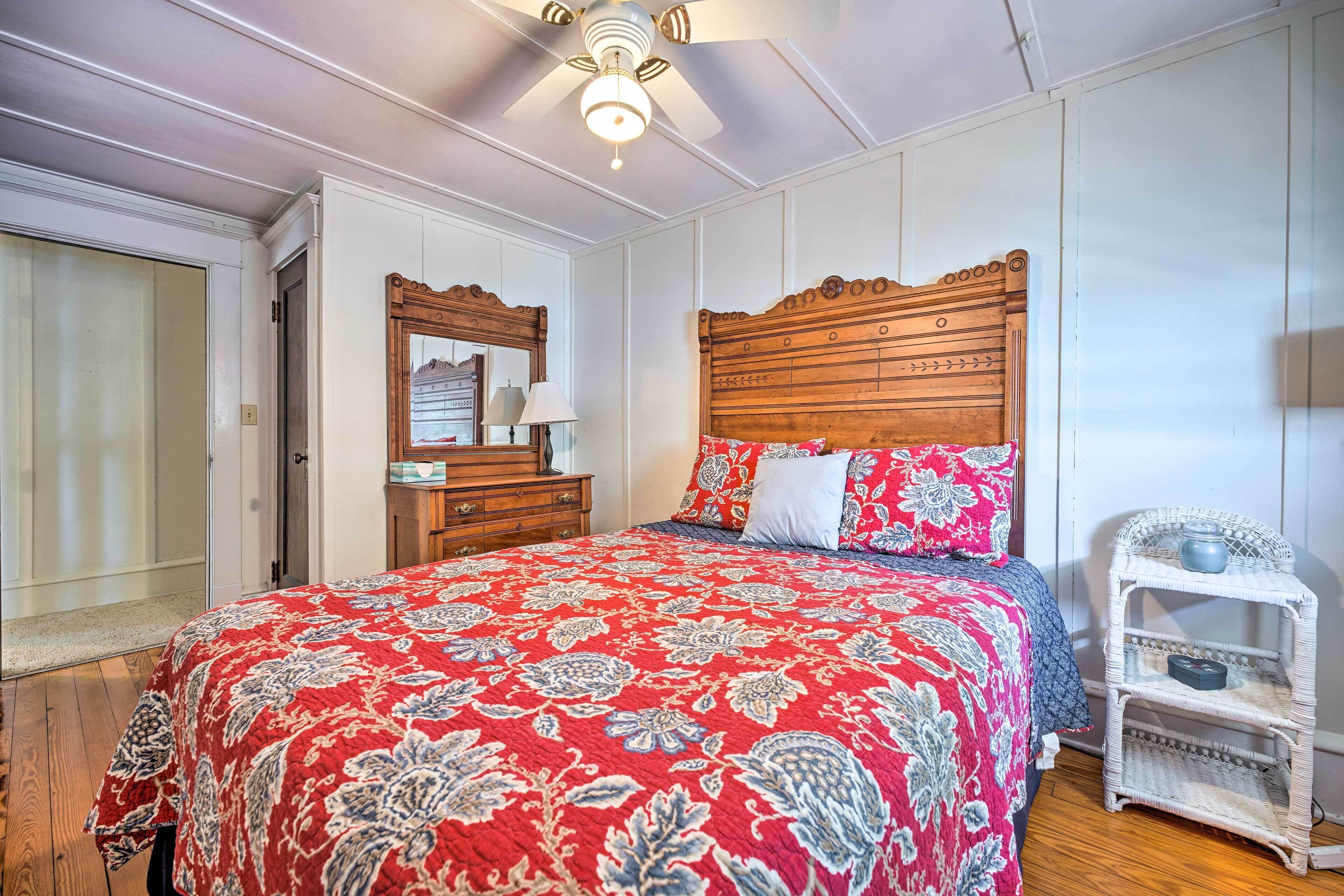 A bedroom with a wooden headboard, a red floral bedspread, a white nightstand with a blue bowl, and a wooden dresser with a mirror.
