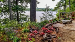 A lakeside garden with a wooden deck, surrounded by trees and various plants. - Lakefront Cottage: Boat Dock, Patio & Kayaks! (Oxford)