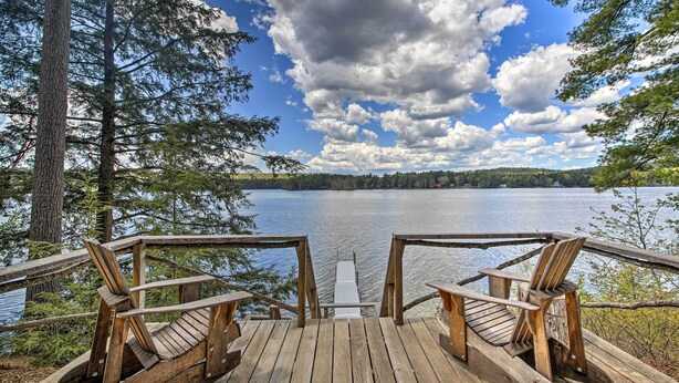 A wooden deck with two Adirondack chairs facing a calm lake, surrounded by trees and a clear sky. - Lakefront Cottage: Boat Dock, Patio & Kayaks! (Oxford)