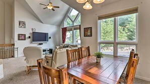 A dining area with a wooden table and chairs, a ceiling fan, a TV mounted on the wall, and large windows with a view of trees. - Lake Harmony Villa: Walk to Big Boulder Mountain! (Lake Harmony)