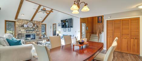 A spacious living room with a large wooden dining table, comfortable seating, a stone fireplace, and a television mounted on the wall.